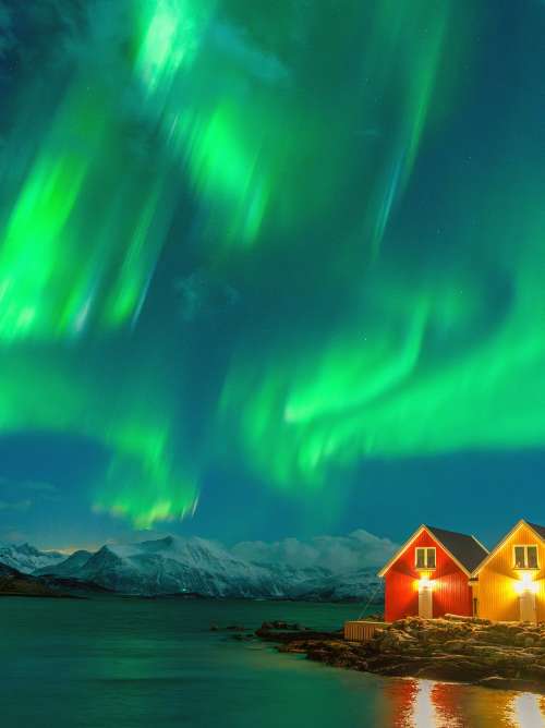 Green lights of Aurora Borealis over snowcapped mountains and cabins by the sea, Sommaroy, Troms og Finnmark, Norway