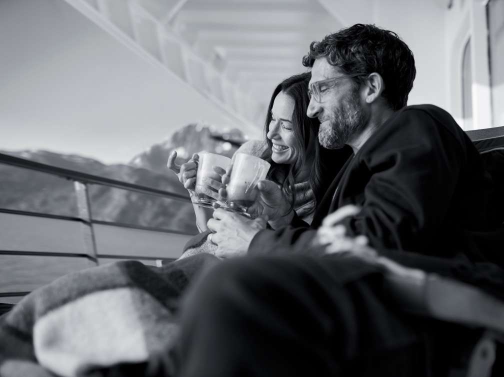 A man and a woman cosy up under a blanket with hot chocolate on the deck of Queen Elizabeth