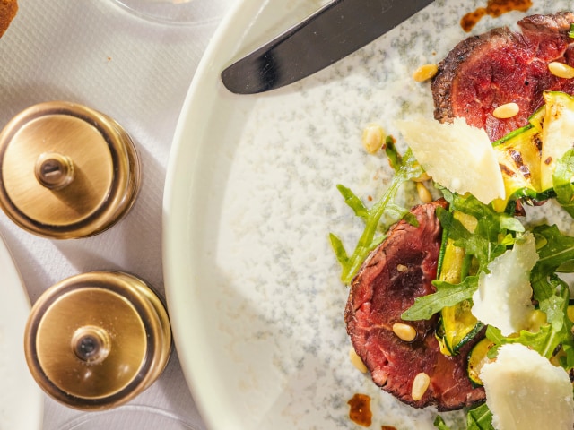Top-down view of a Cunard dining table, including golden salt and pepper mills, a beef entree, a tomato salad and freshly baked bread rolls, accompanied with white wine