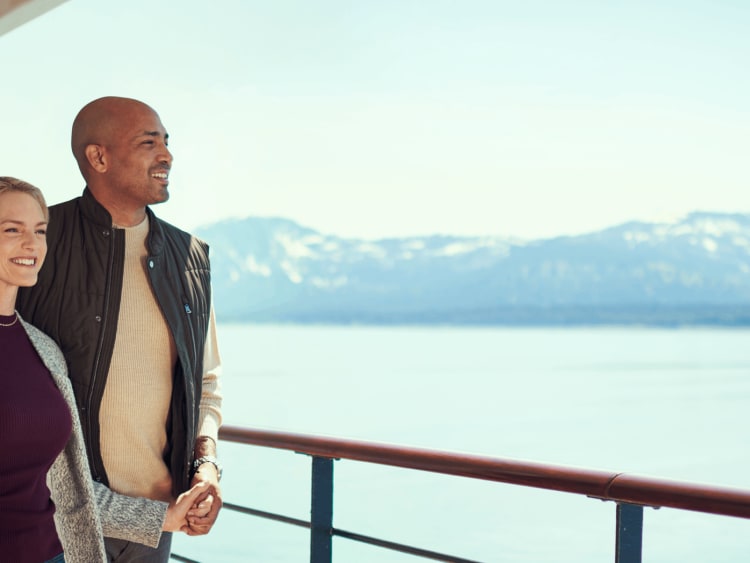Couple enjoying scenic ocean views from the deck of a luxury cruise ship with snow-capped mountains in the distance