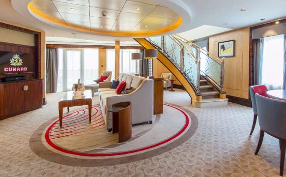Lower floor living area of a Q1 Grand Duplex Queens Grill Suite on Queen Mary 2, showing sweeping staircase, sofa, wall-mounted television, balcony access and dining area