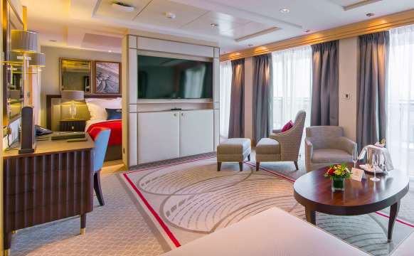 A typical Q4 Penthouse Queens Grill Suite on Queen Mary 2, showing kingsize bed behind room partition, wall-mounted television, comfortable living area, desk and balcony access