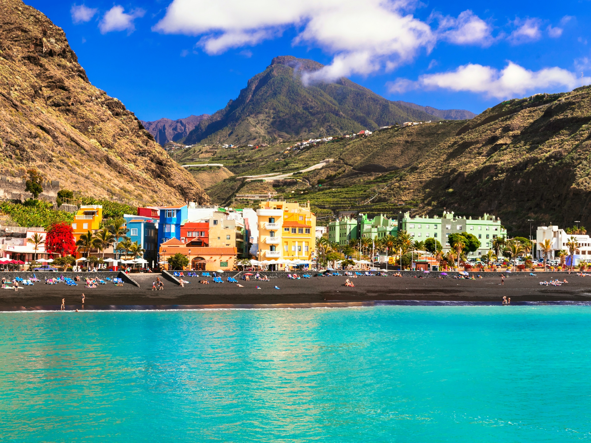 A daytime, eye-level shot of a coastal scene in La Palma, Spain, featuring vibrant turquoise-blue water in the foreground. A wide dark sand beach stretches behind the water, lined with a row of brightly painted colorful buildings. Towering, steep green and brown mountains rise dramatically behind the town under a bright blue sky dotted with white clouds.