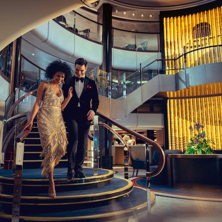 A man and a woman in eveningwear descend the staircase in the Grand Lobby on a Queen Anne Gala Evening