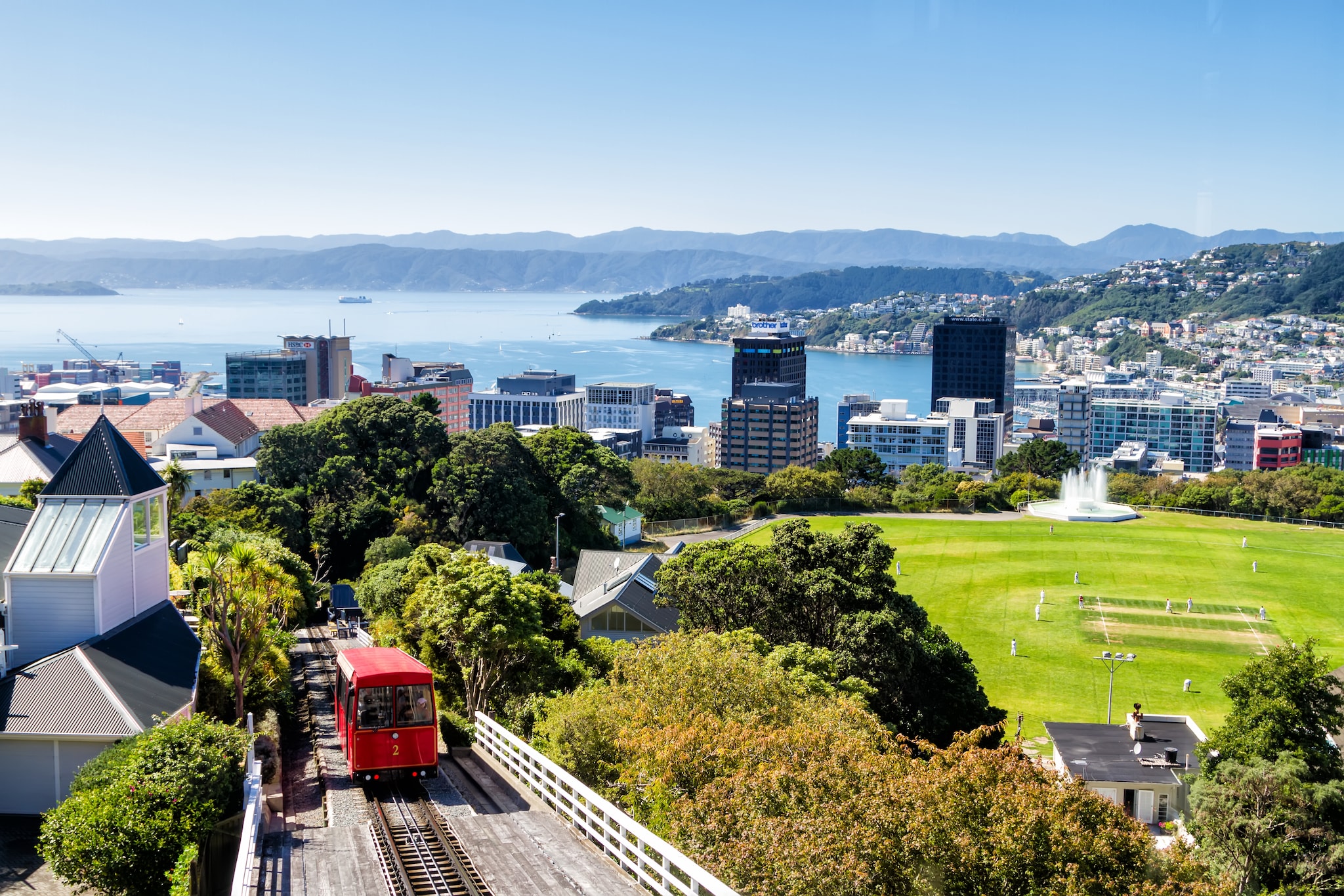 Red cable car and sports field above downtown Wellington, bay and mountains beyond